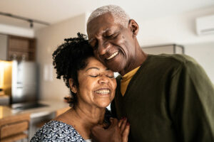 A husband who is a spousal caregiver hugs his wife as they smile.