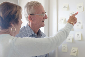 A woman points to notes posted on the wall, one way to improve dementia care, as her elderly father looks on.