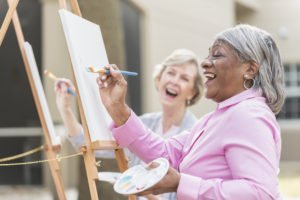 Two multi-ethnic senior women sitting outdoors at easels painting pictures on canvases. The focus is on the African American woman who is holding a paintbrush and looking up at her artwork as she laughs.